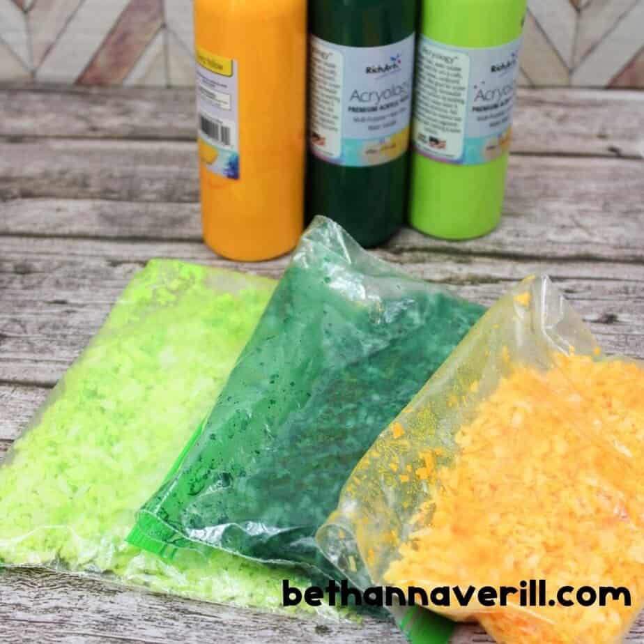 Three zip-top bags containing dyed coconut in light green, dark green, and orange are laid out in front of coordinating paint bottles, ready to dry or mix.