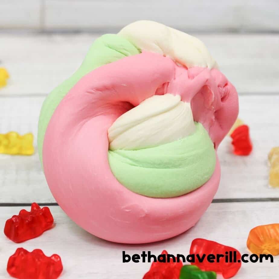 A twisted swirl of creamy pink, mint green, and white edible slime rests on a white wood surface, surrounded by gummy bears in various colors. The soft, stretchy texture is highlighted.