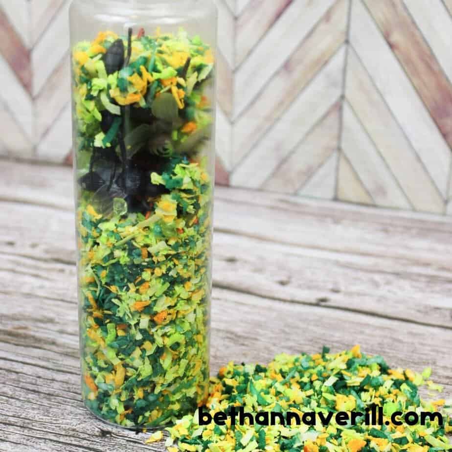 A clear bottle stands upright, stuffed with dyed coconut flakes and hidden toy insects. A pile of excess green and yellow flakes is scattered at the base.