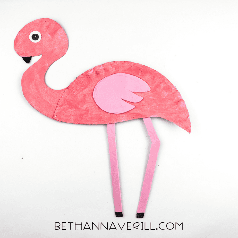 Finished pink flamingo paper plate craft with a cut-out wing, black beak, and black-tipped legs, laying flat on a white background.