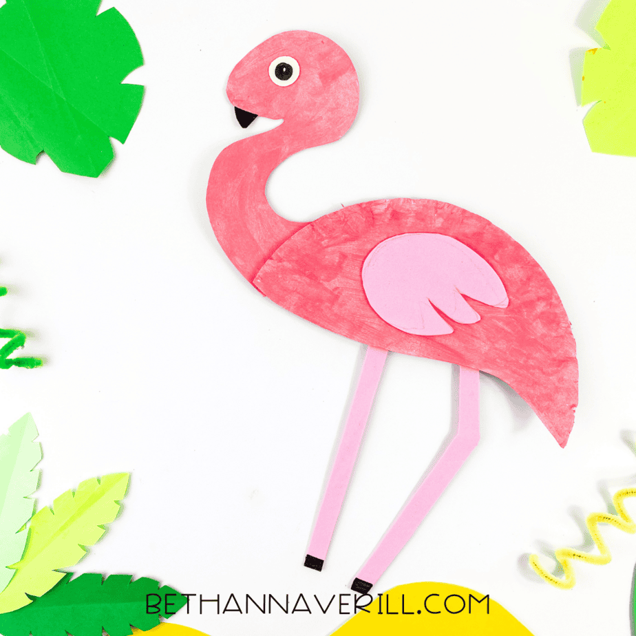 Flamingo paper plate craft displayed with colorful paper leaves and curly pipe cleaners, creating a tropical scene on a white surface.