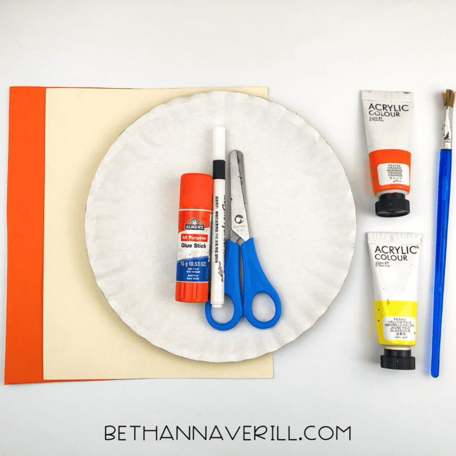Overhead view of craft supplies: paper plate, glue stick, scissors, black marker, orange and cream construction paper, orange and yellow paint, and a paintbrush.