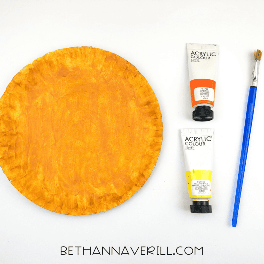 A paper plate painted a golden orange color, with orange and yellow paint tubes and a paintbrush beside it.