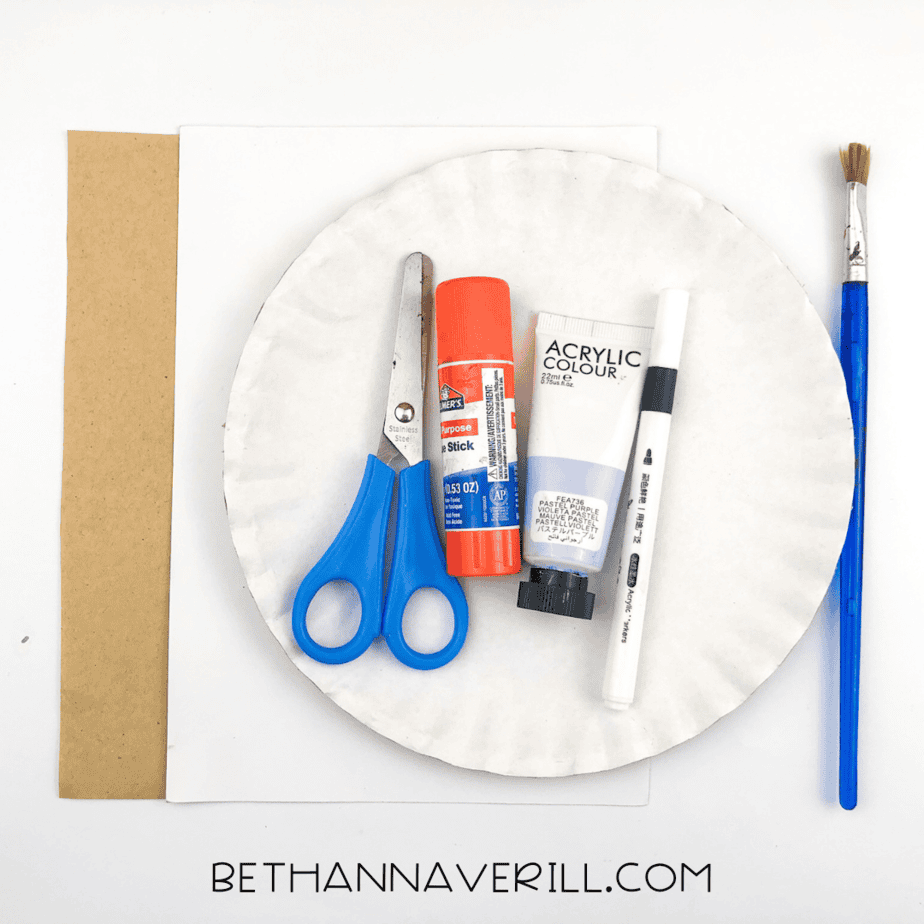 Craft supplies laid out for a hippo paper plate craft, including scissors, glue stick, purple paint, a paintbrush, a black marker, a white paper plate, and brown and white paper.