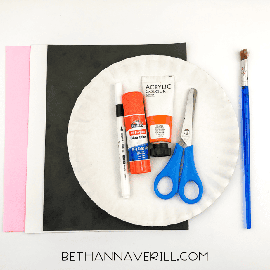Craft materials laid out on a table, including paper plate, paint, glue stick, scissors, marker, and colored paper