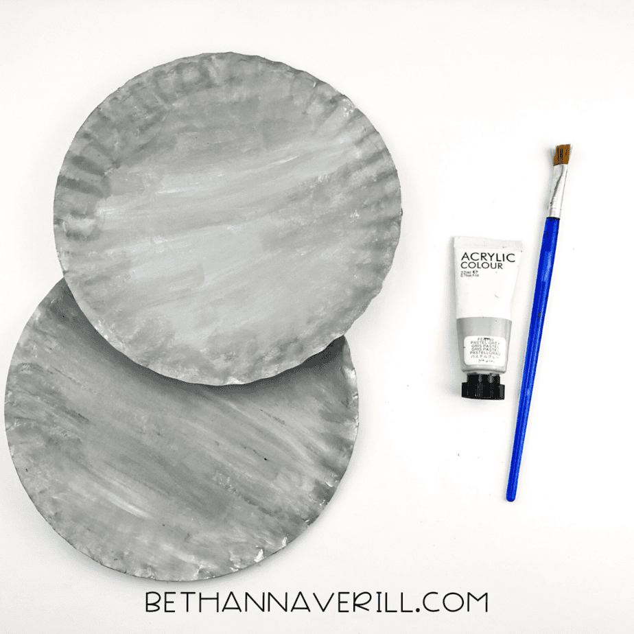 Two paper plates fully painted gray with a paintbrush and gray acrylic paint beside them.