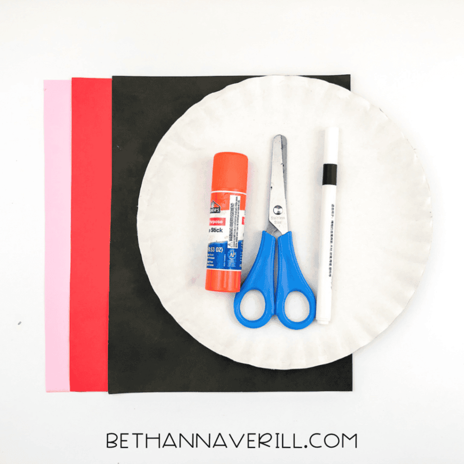 A paper plate, glue stick, kid scissors, black marker, and sheets of black, red, and pink construction paper arranged on a white background.