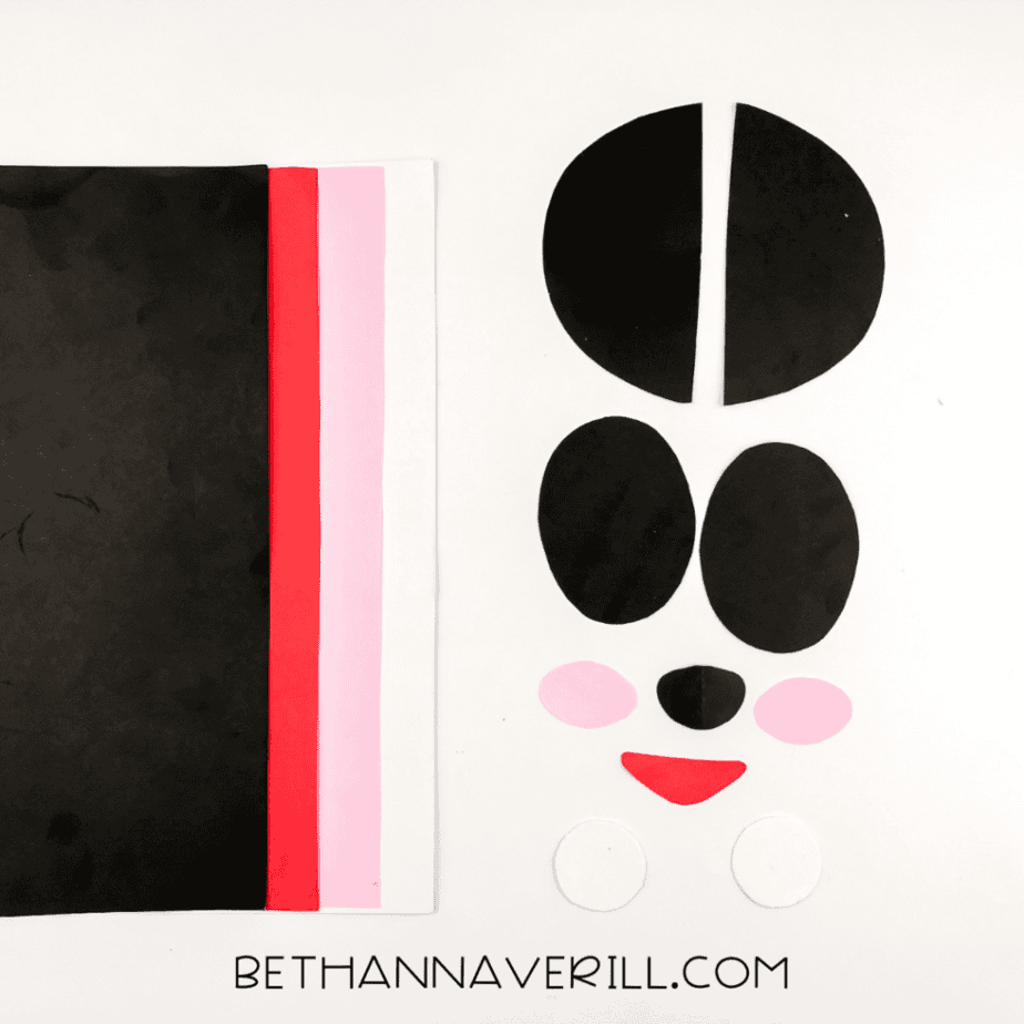Construction paper pieces cut into shapes for ears, eye patches, cheeks, eyes, nose, and a red mouth, laid out next to the paper sheets.