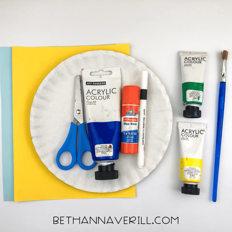 A paper plate surrounded by blue, green, and yellow acrylic paint, glue stick, scissors, paintbrush, black marker, and blue and yellow construction paper.