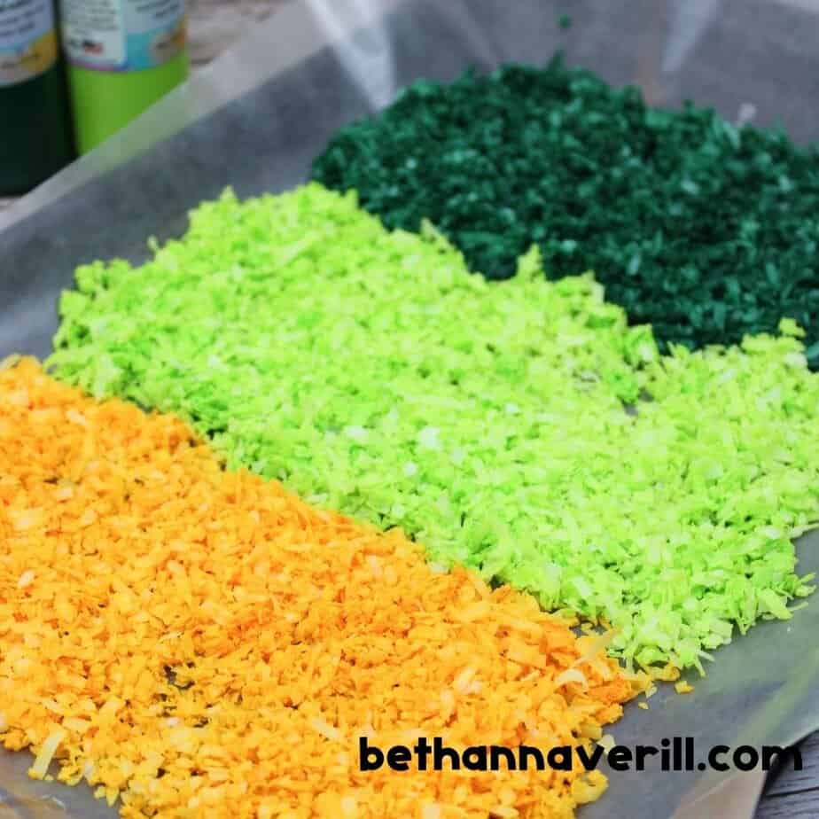 Four neat piles of dyed coconut shreds in dark green, light green, bright yellow, and orange are arranged on wax paper, with bottles of acrylic paint in the background.