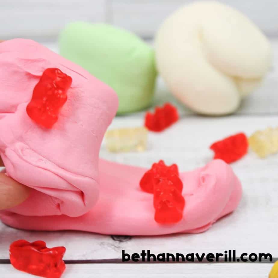A close-up of stretchy pink slime with red gummy bears pressed into it, being pulled by a hand. In the background are pastel balls of green and white slime.