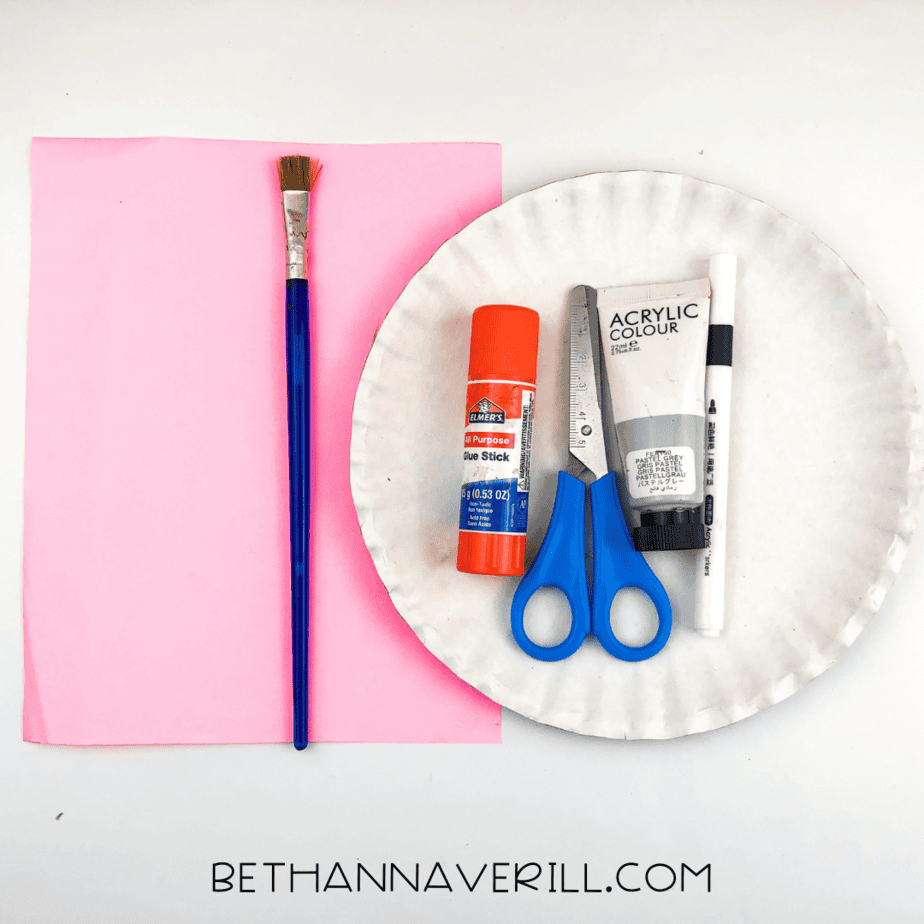 A flat lay photo showing a paper plate, pink construction paper, gray acrylic paint, paintbrush, scissors, glue stick, and black marker.