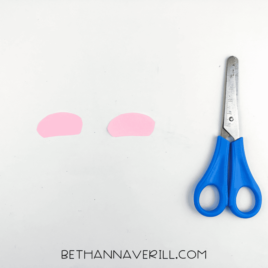 Two small pink paper ovals placed next to a pair of scissors, ready to be glued as inner ears.