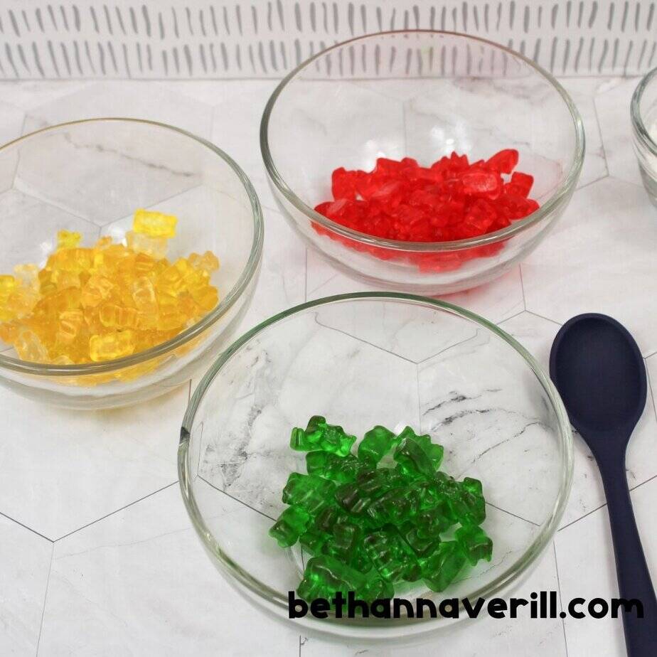 Small glass bowls each hold gummy bears sorted by color—yellow, red, and green—with a navy spoon nearby, ready for making multi-colored slime.