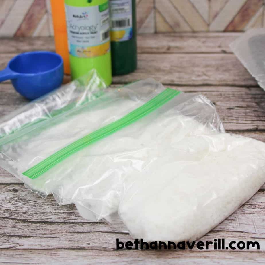 Two plastic zip-top bags filled with plain white coconut flakes sit next to bottles of green and yellow acrylic paint and a blue measuring cup, all on a rustic wooden surface.