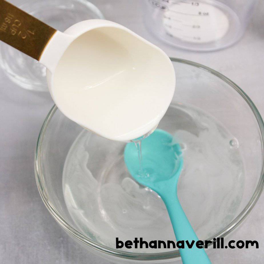 A half-cup of white glue is being poured into a bowl with clear liquid, stirred by a turquoise spoon. Measuring tools and bowls are visible in the background.