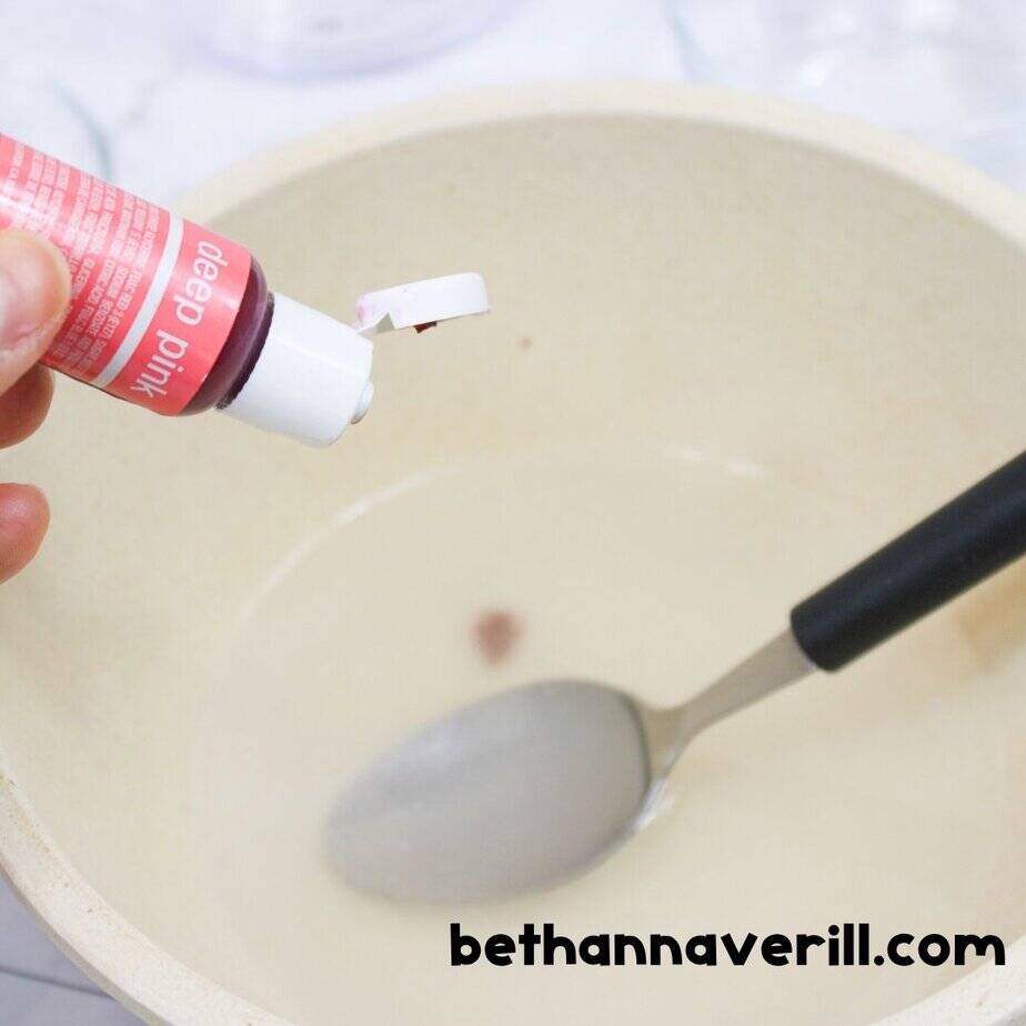 A bottle of deep pink food coloring being added to a bowl of slime mixture to color unicorn slime, with spoon inside the bowl.