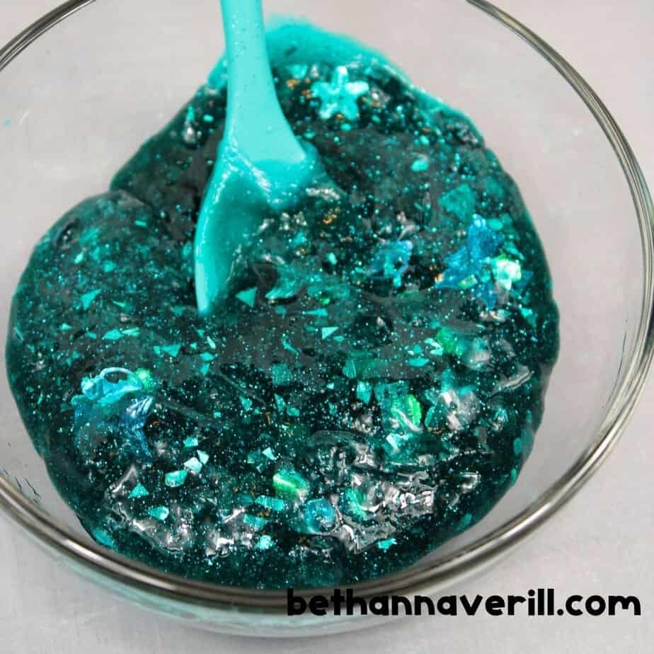 A bowl of deep teal glitter slime is being stirred with a turquoise spoon, showing the slime pulling away from the edges as it thickens. The mixture sparkles with chunky glitter and iridescent shapes.