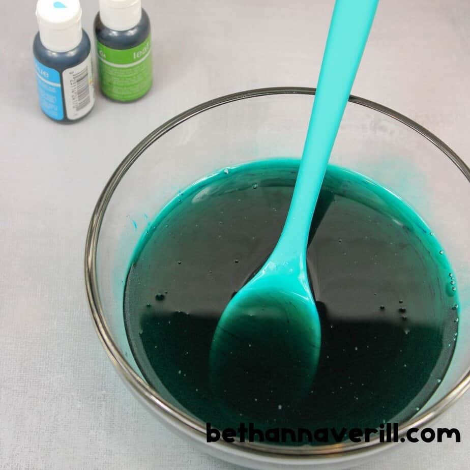 A bowl filled with dark teal liquid slime base is being stirred with a turquoise spoon, next to green and blue bottles of food coloring labeled “leaf green” and “sky blue.”