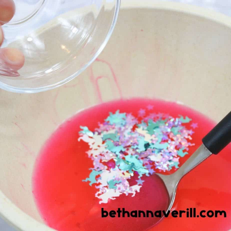 Glitter and unicorn-shaped confetti added to pink liquid in a mixing bowl as part of a fun unicorn slime recipe for kids.