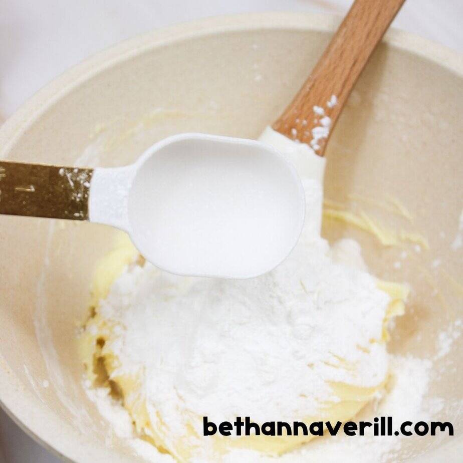 Close-up of a measuring cup pouring cornstarch onto a mixture of vanilla pudding and slime ingredients in a beige bowl, with a spatula in the mixture. bethannaverill.com branding visible.