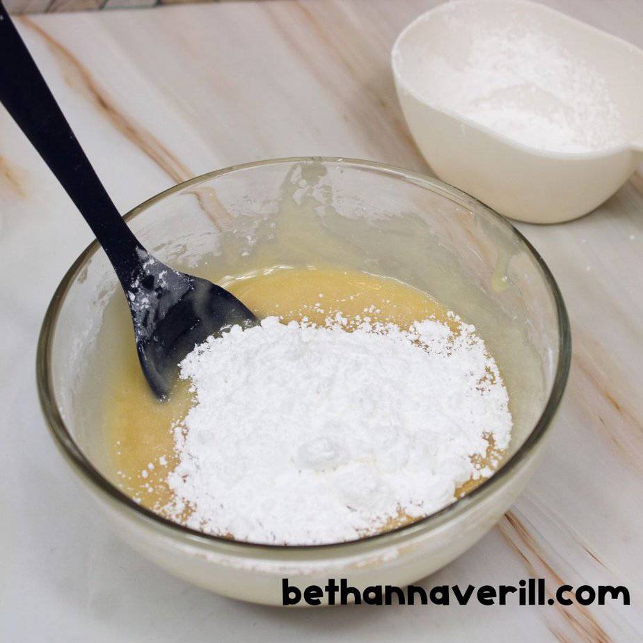 A mixing bowl of honey slime with a generous pile of powdered sugar on top, being stirred with a black spoon. A second container of sugar sits nearby.