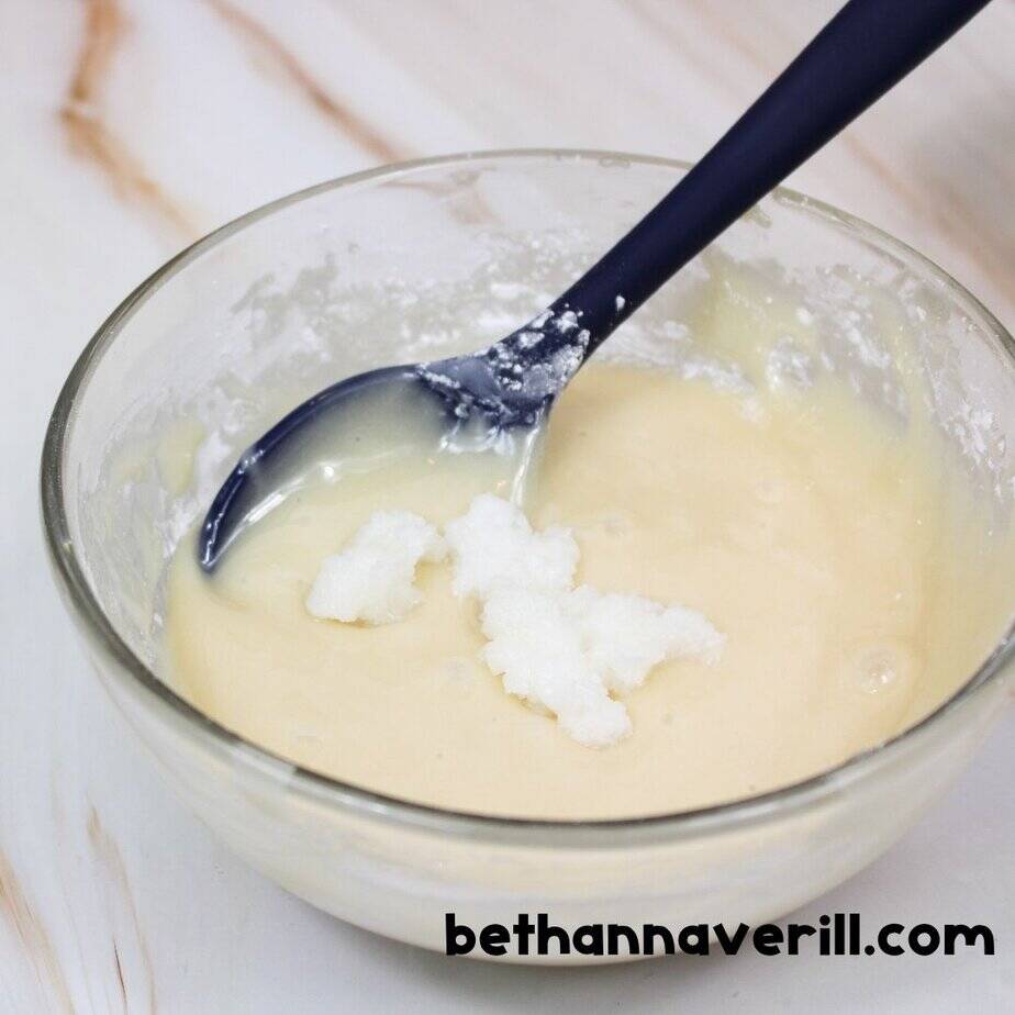 A glass bowl of honey slime batter with white coconut oil scooped on top, mid-recipe, and a blue spoon resting inside the bowl. bethannaverill.com is at the bottom.