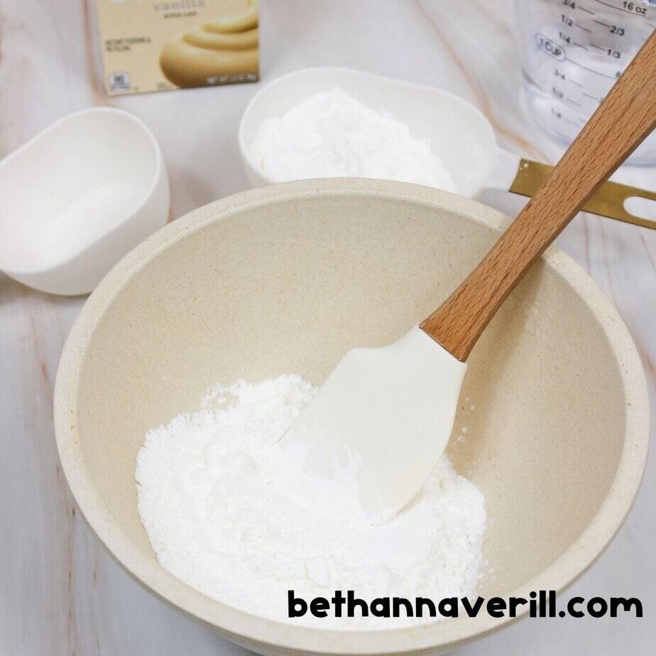 Beige mixing bowl with a white spatula stirring dry ingredients, including cornstarch and pudding mix. Additional measuring cups of sugar and cornstarch are in the background. bethannaverill.com branding shown.
