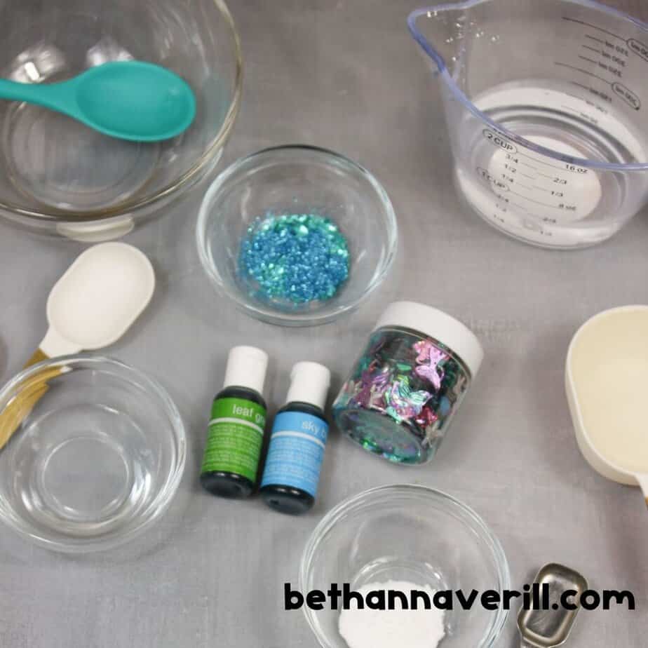 A flat lay of all the supplies needed for mermaid slime: clear glue, food coloring, measuring cups, bowls, glitter, and a jar of assorted pastel sequins in mermaid shapes like tails and stars.