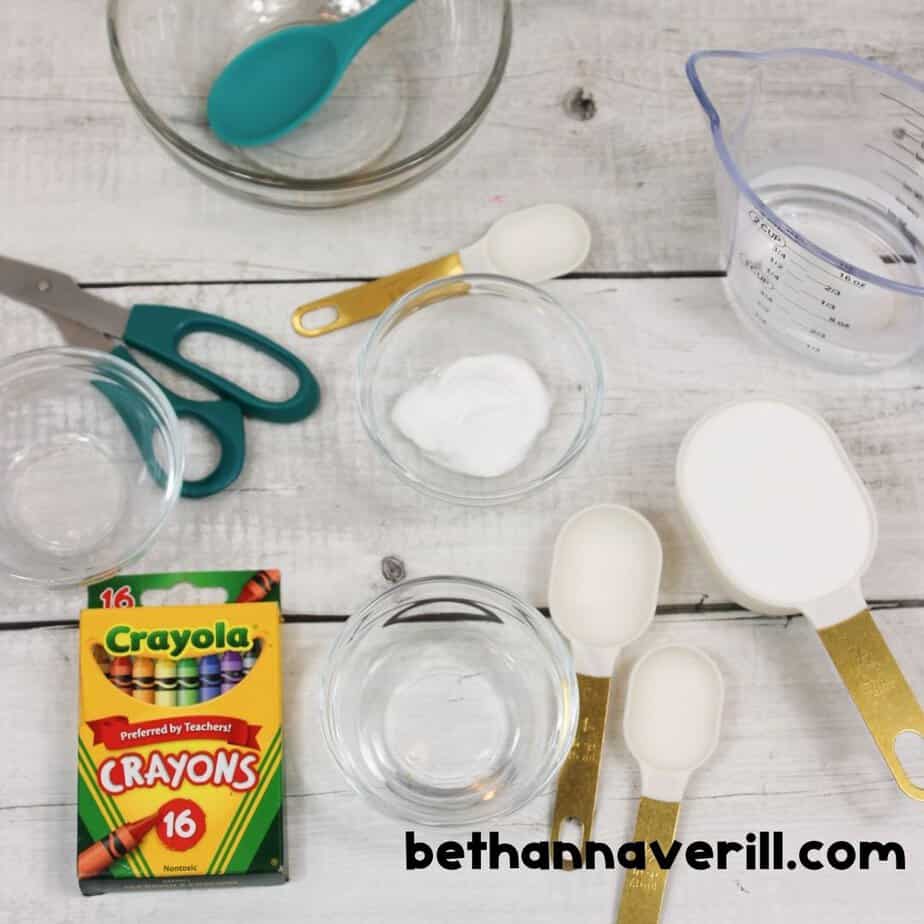 Flat lay of ingredients and tools for making crayon slime, including glue, baking soda, water, crayons, measuring spoons, and bowls.