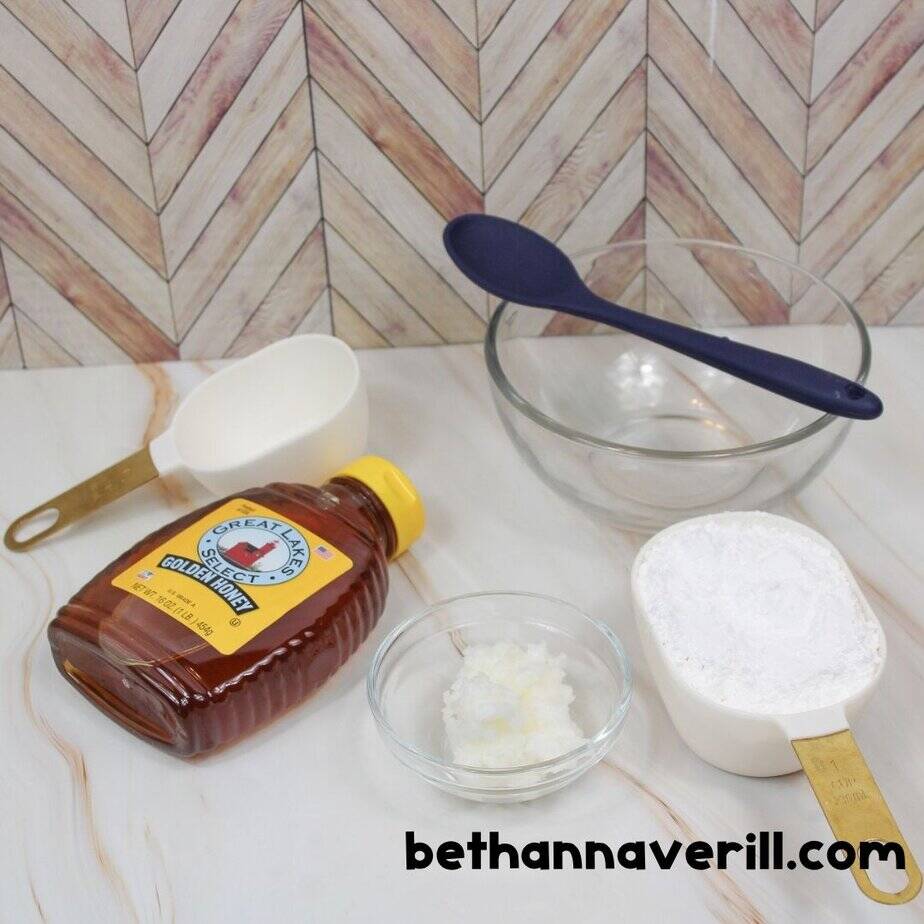 Honey slime ingredients displayed on a countertop: a bottle of Great Lakes Select Golden Honey, a measuring cup of powdered sugar, a small dish of coconut oil, an empty measuring cup, a glass mixing bowl, and a blue spoon. bethannaverill.com is printed at the bottom.