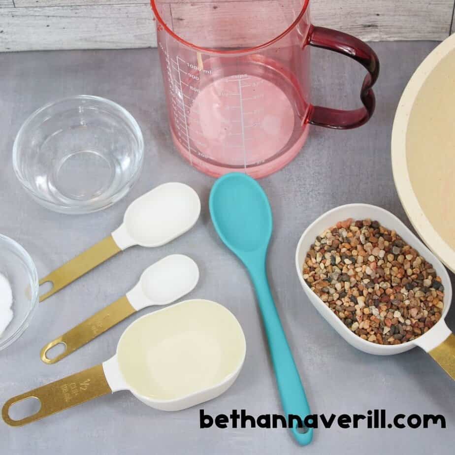 A flat lay of slime-making supplies for pebble slime, including a red measuring pitcher, teal spoon, measuring cups, a small bowl of baking soda, and a container of small pebbles.