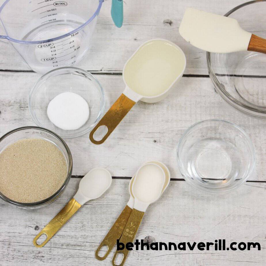 A flat lay of ingredients and tools for sand slime including measuring cups, a spatula, clear glue, baking soda, water, and a bowl of sand.