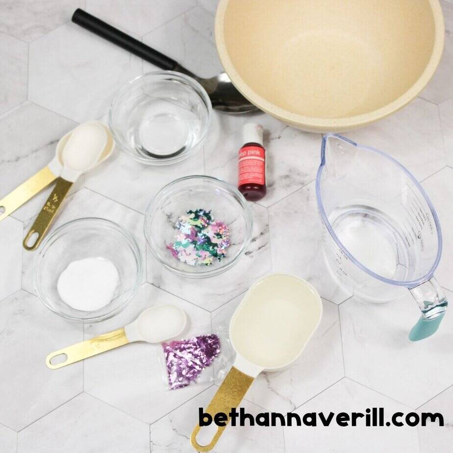 Top-down view of unicorn slime ingredients including glitter, food coloring, measuring cups, contact solution, and a mixing bowl.