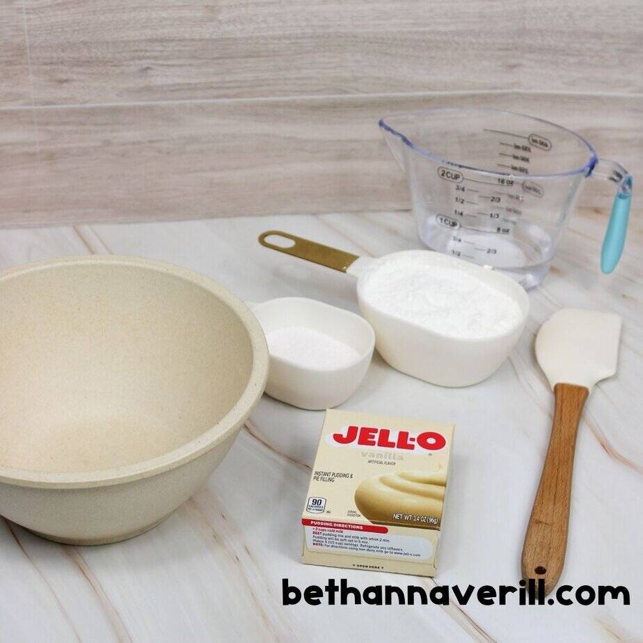 Ingredients and supplies for making vanilla pudding slime, including a mixing bowl, Jello vanilla pudding mix box, measuring cups with cornstarch and sugar, water in a measuring cup, and a spatula on a marble countertop. bethannaverill.com branding is visible.