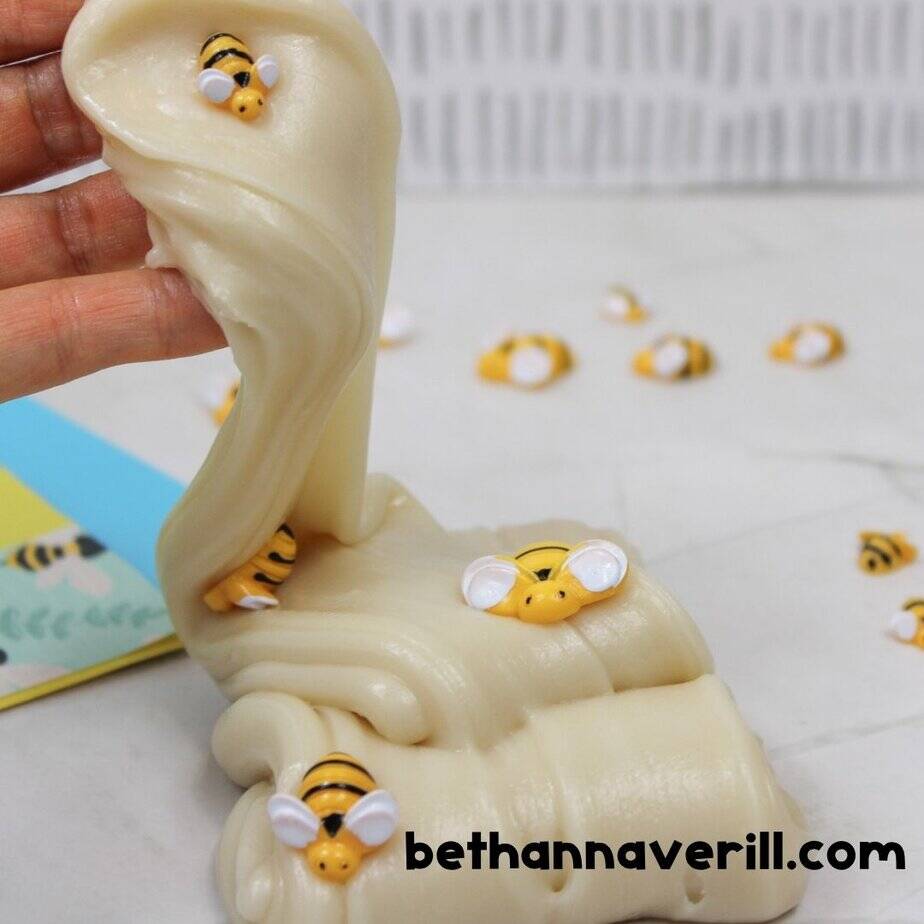 Glossy honey-colored slime being pulled upward by hand, decorated with cute plastic bees, with more bee embellishments scattered on the surface. bethannaverill.com is shown at the bottom.