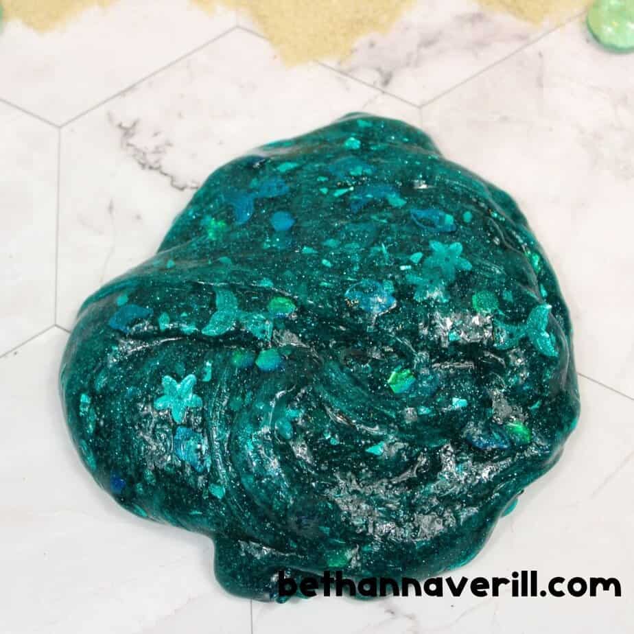 A finished lump of shiny, dark teal mermaid slime rests on a marble surface, filled with glitter and mermaid-themed sequins like tails and flowers.