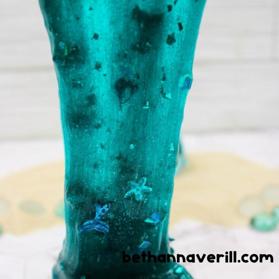 A close-up of stretchy teal mermaid slime being pulled upward, packed with glitter and sea-themed sequins like mermaid tails and stars.