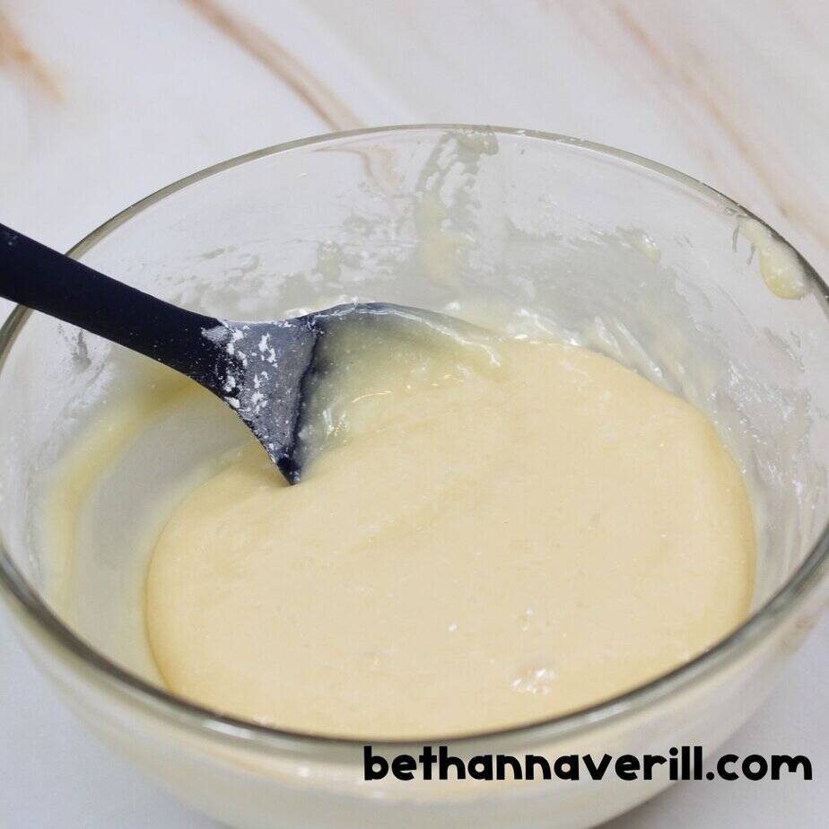 Thick mixture of honey-colored slime in a glass bowl being stirred with a dark blue spatula, partially blended and still gooey.