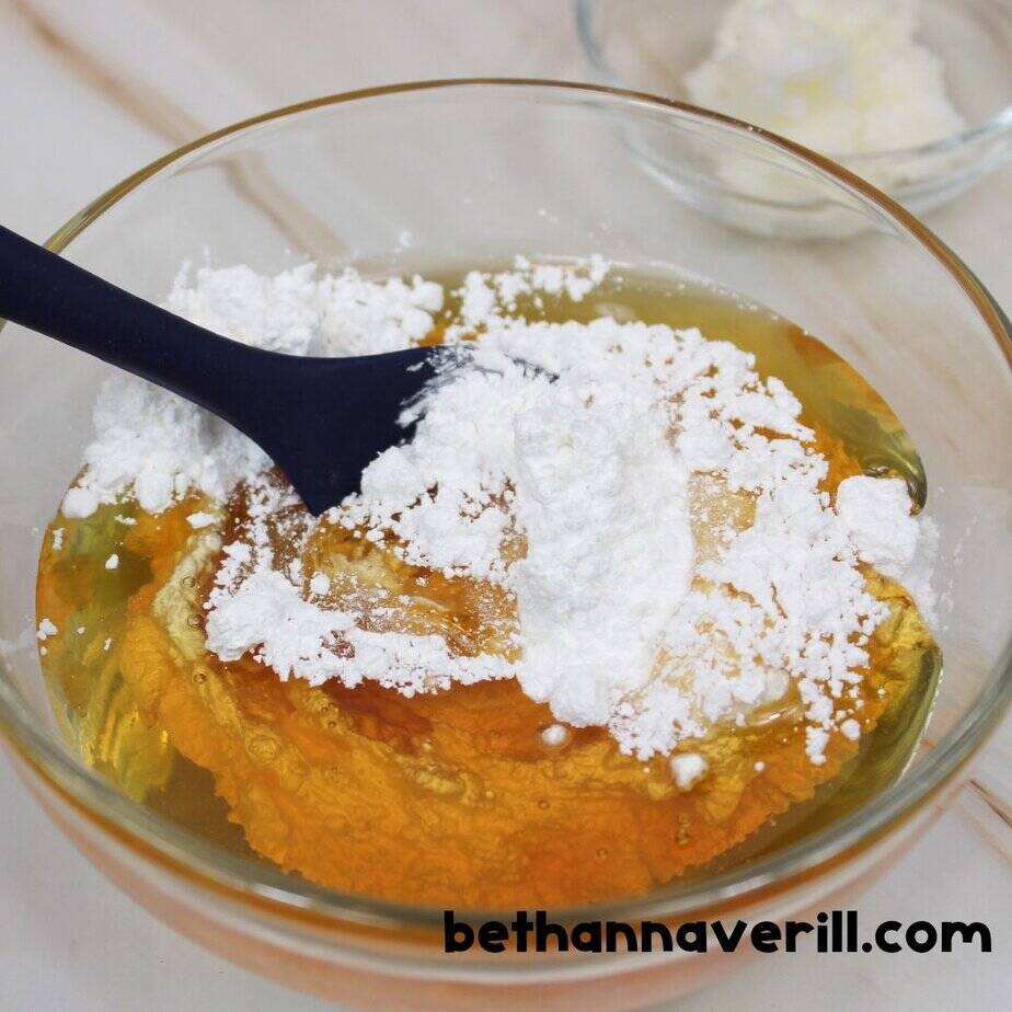 Unmixed honey and white powdered sugar in a glass bowl with a blue spatula, ready to be stirred; a small dish of white solid ingredient is visible in the background.