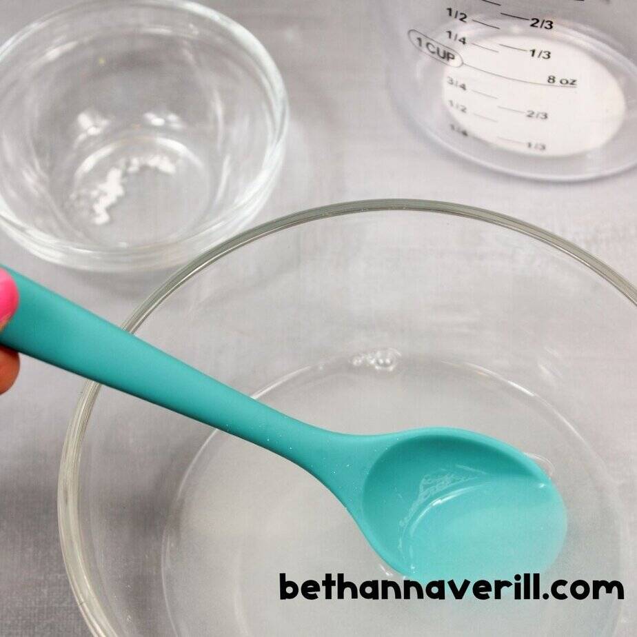 A hand with pink nail polish holds a turquoise spoon in a bowl of clear liquid, next to a measuring cup and small glass bowl of baking soda.