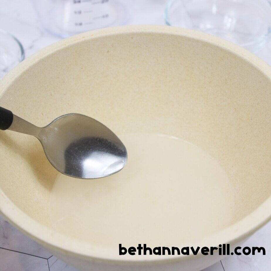 Mixing spoon resting in a bowl of clear liquid, showing the start of a unicorn slime recipe with water and baking soda.