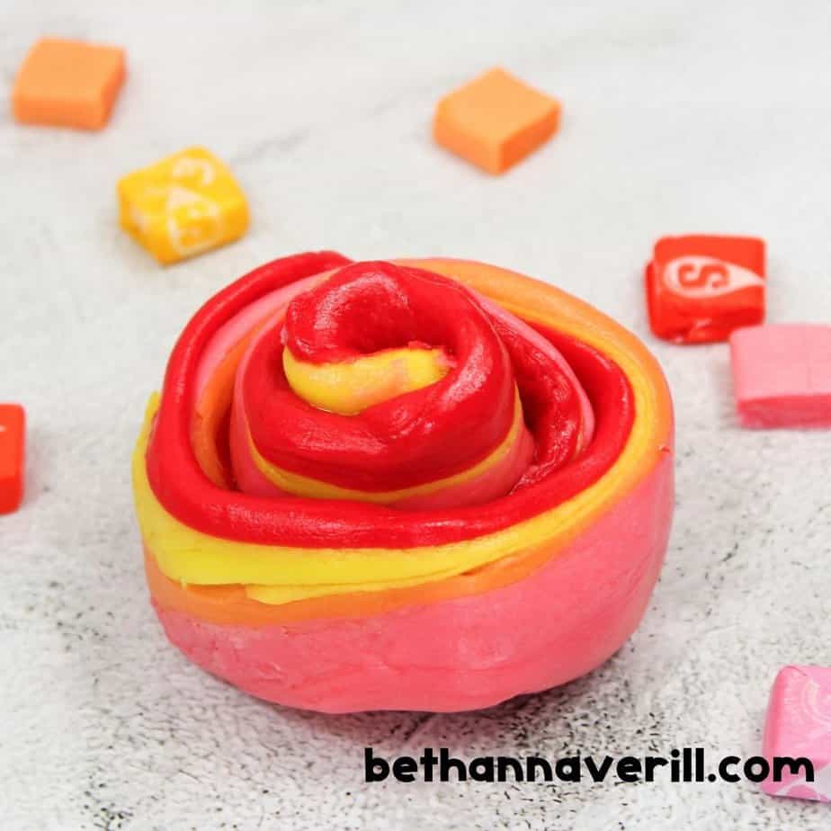 A colorful, twisted rose-like swirl of Starburst slime in red, orange, yellow, and pink sits on a speckled countertop, with unwrapped and whole Starburst candies scattered in the background.