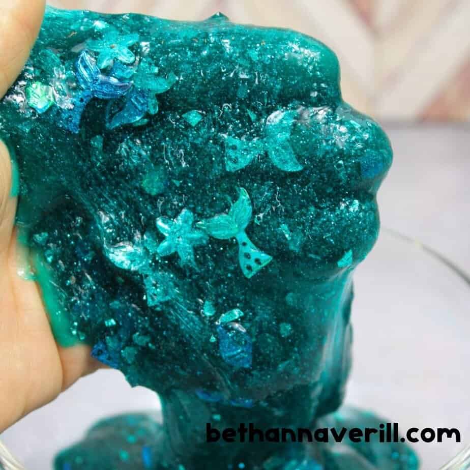 A close-up of someone holding stretchy mermaid slime, sparkling with blue and teal glitter and packed with sequins shaped like mermaid tails and flowers.v