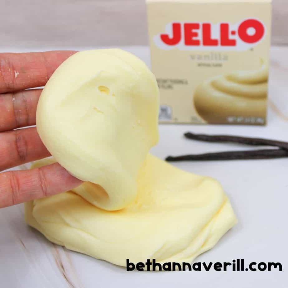 Hand lifting a stretchy, smooth piece of vanilla pudding slime from a marble surface, with the Jello vanilla pudding box and vanilla beans in the background. bethannaverill.com branding shown.