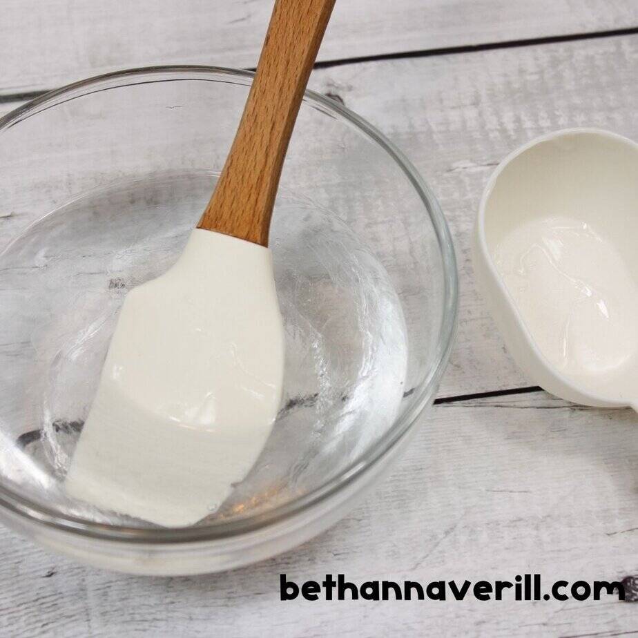 A white rubber spatula rests in a glass bowl coated with clear glue to make spring slime, with a white measuring cup nearby that held the glue.