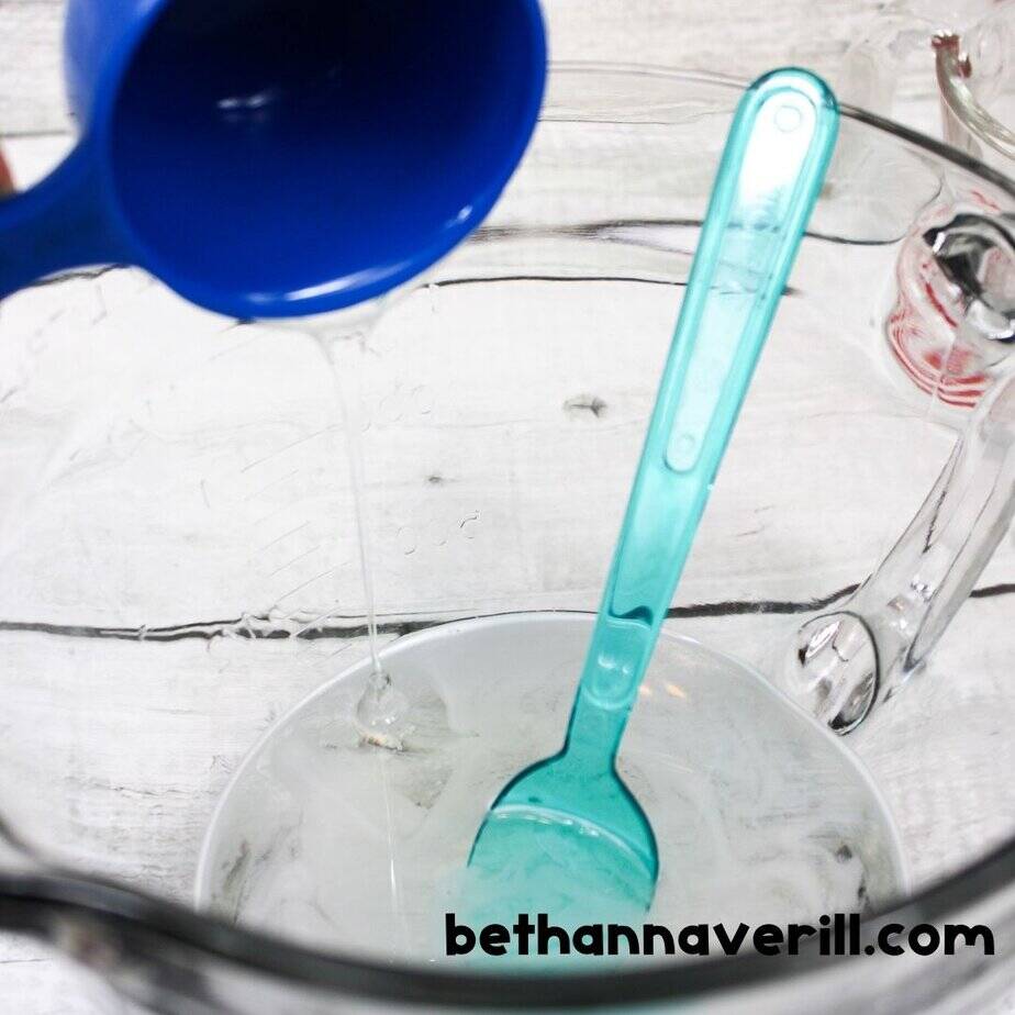 Clear glue is being poured from a blue measuring cup into a large glass mixing bowl, which already contains some glue and a teal plastic spoon.
