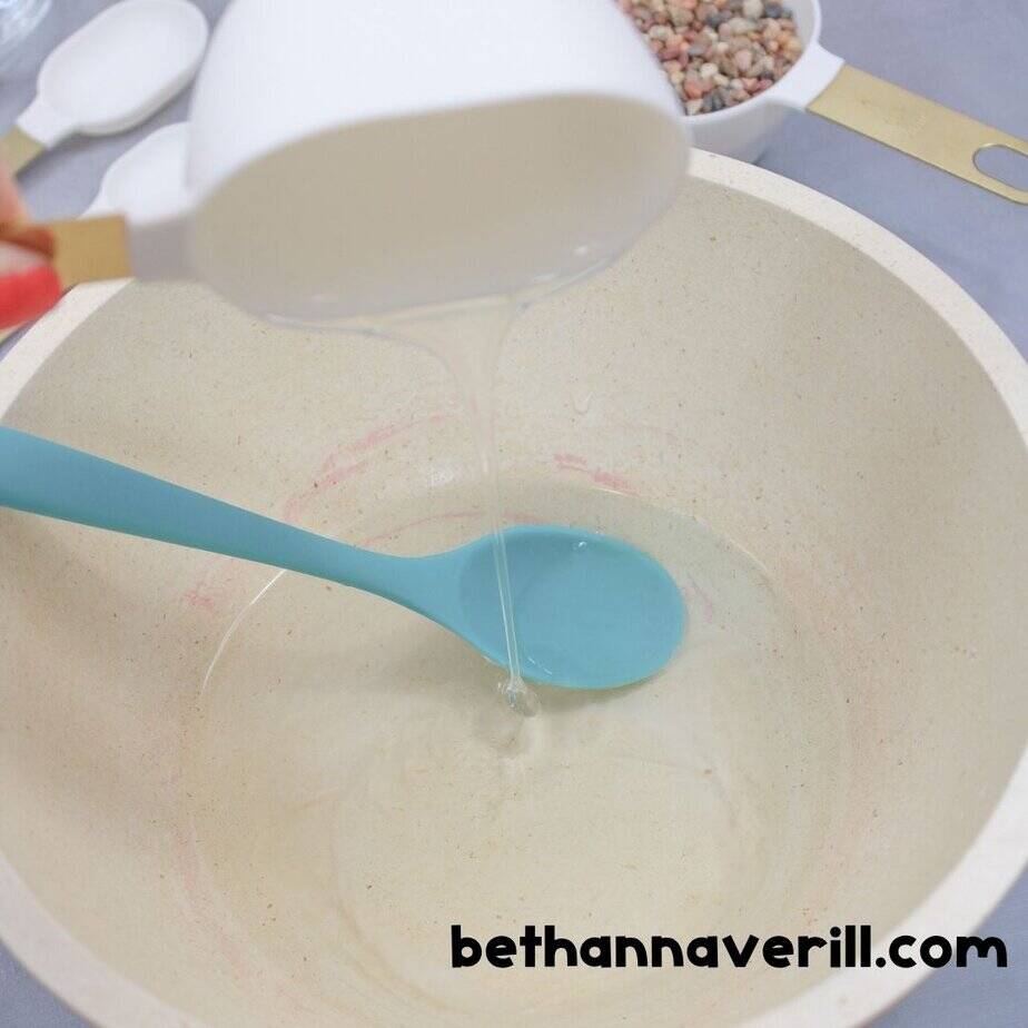 A close-up of clear glue being poured from a white measuring cup into a large cream-colored mixing bowl with a teal spoon inside. The glue forms a thin stream as it enters the bowl.