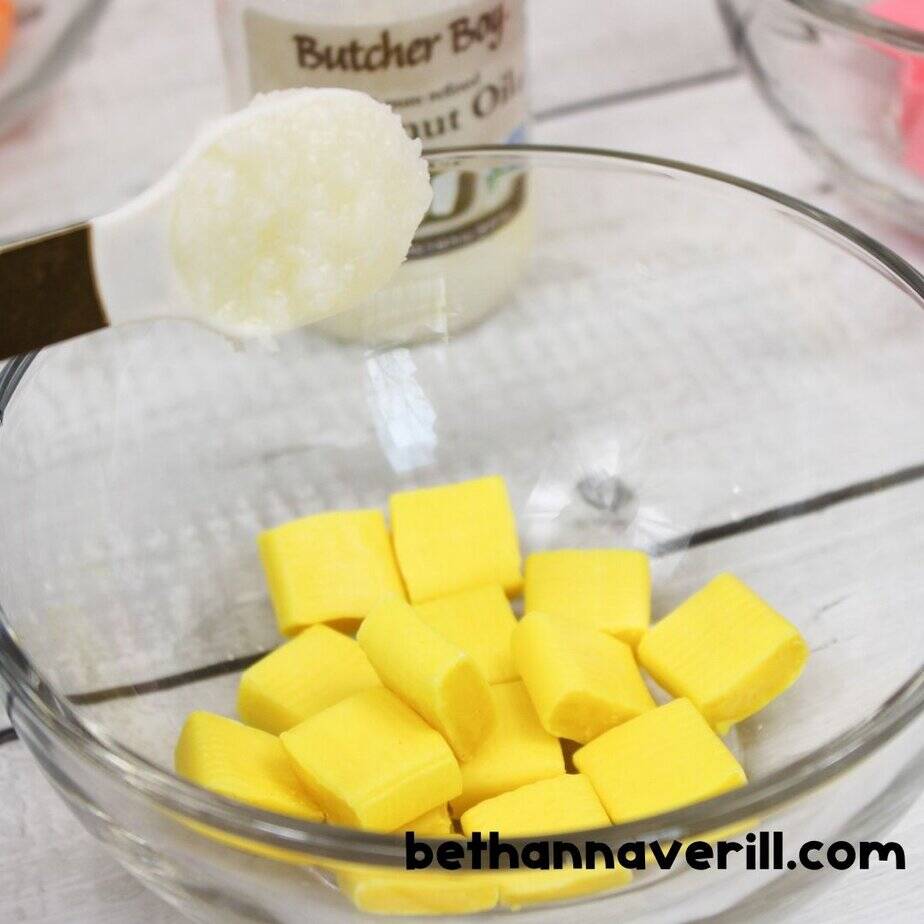 Glass bowl with yellow Starburst candies and a spoonful of coconut oil being added, ready to melt for slime making.