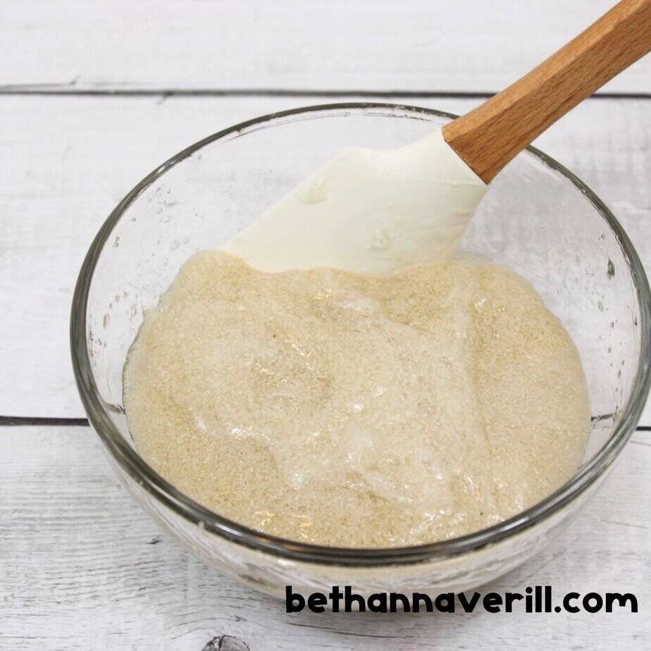 A rubber spatula mixes fine tan sand into clear slime in a glass bowl, giving the slime a textured, grainy appearance.